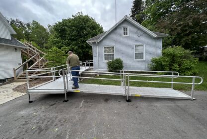 A man stands on a metal accessibility ramp leading to the entrance of a small white house surrounded by trees and shrubs.