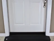 White door with a silver handle and a black draft stopper at the bottom, set against a beige wall and light-colored floor.