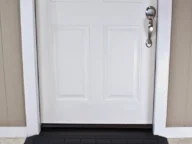 White door with a silver handle, surrounded by a light brown wall panel. A black rectangular mat lies centered in front of the door.