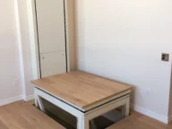 A residential vertical platform lift with a wooden platform and glass panel, installed in a room with light wood flooring and white walls.