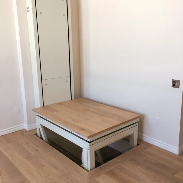 A residential vertical platform lift with a wooden platform and glass panel, installed in a room with light wood flooring and white walls.
