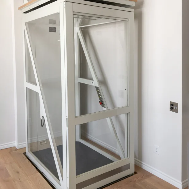 Glass residential elevator with metal frame installed indoors, featuring a control panel inside and the Savaria logo in the lower left corner.