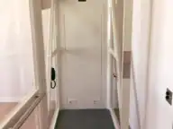 A small residential elevator with glass walls, a gray floor, and control panel, inside a house with light-colored wooden floors.