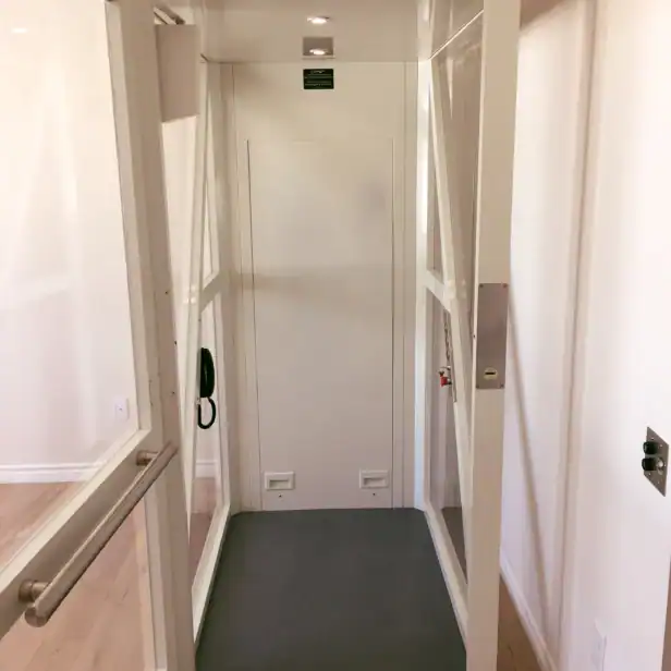 A small residential elevator with glass walls, a gray floor, and control panel, inside a house with light-colored wooden floors.