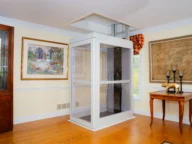 A small white residential elevator is installed in the corner of a room with wooden floor, surrounded by framed artwork and furniture.