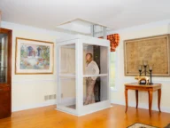 A woman stands inside a home elevator positioned in a living room, surrounded by framed artwork, wooden furniture, and light-colored walls.