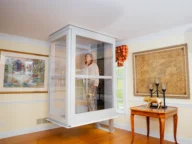 A woman stands inside a transparent residential elevator cabin installed in a living room with framed artwork on the walls and wooden furniture.
