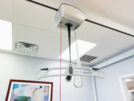 Ceiling-mounted patient lift with a red laser guide and control panel. A framed abstract painting is visible on the wall in the background.