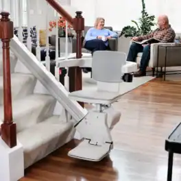 Bruno Elan SRE-5000 stairlift parked at the bottom of a carpeted staircase while a couple talks in the living room