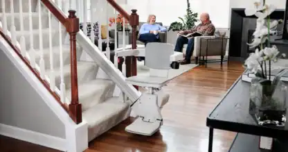 Bruno Elan SRE-5000 stairlift parked at the bottom of a carpeted staircase while a couple talks in the living room