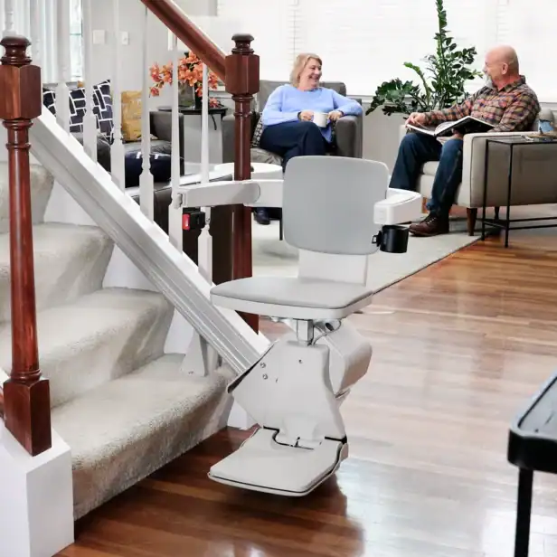 Bruno Elan SRE-5000 stairlift parked at the bottom of a carpeted staircase while a couple talks in the living room