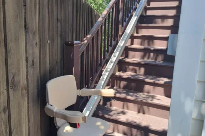 A brown stairlift installed beside a wooden staircase, next to a tall wooden fence, with trees visible in the background.