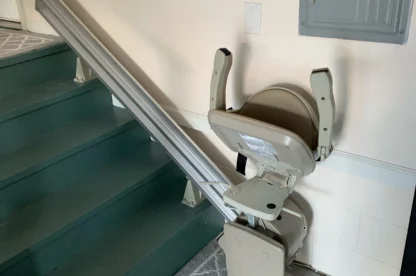 A stairlift is installed next to a green staircase, with the chair positioned at the bottom. The wall and a control panel are visible in the background, and a gray carpet lies on the floor.