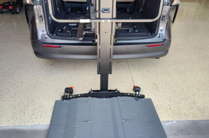 A van with its rear hatch open, showing a deployed wheelchair lift platform extending onto the garage floor.