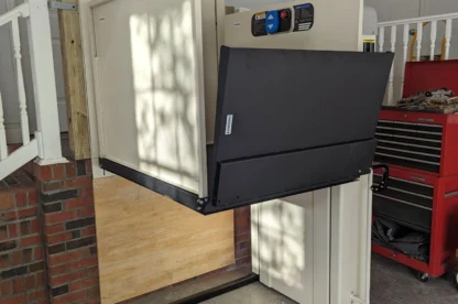 A stairlift installed next to a short flight of stairs, featuring a fold-out platform for wheelchair access, with a brick and wood wall in the background and a red tool cabinet nearby.