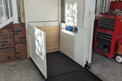 A residential wheelchair lift installed in a garage, featuring a platform, control panel, and safety barriers. Background includes part of a staircase, red tool chest, and concrete floor.