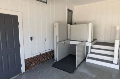 A home entryway shows a wheelchair lift adjacent to a short flight of stairs, facilitating access to the raised doorway. The space has a wall-mounted control panel and a grey-painted door.