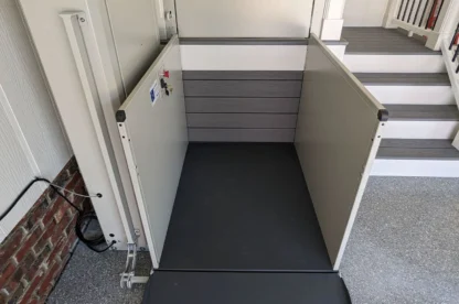 A vertical platform lift next to a set of stairs, featuring a ramp and safety walls, designed to assist with accessibility in a residential setting.