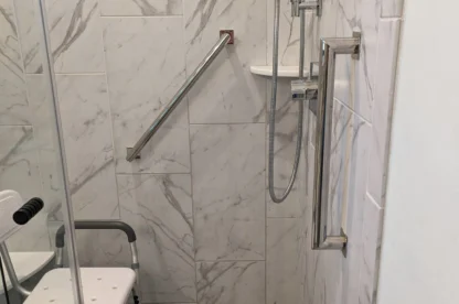 A shower with a marble-tiled wall, equipped with a handheld showerhead, a metal grab bar, a wall-mounted chair, and a non-slip mat on the floor. A glass partition is partially visible.