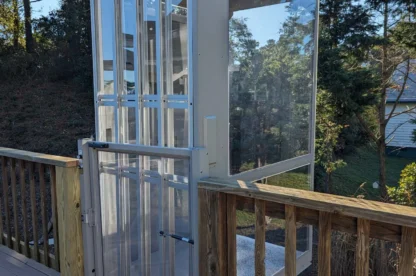 Outdoor glass and metal elevator shaft extending above a wooden deck, surrounded by trees in the background.