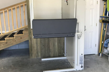 An indoor stairlift installed beside a wooden staircase in a garage, with a folded platform and a white door in the background.