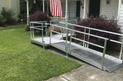 A metallic wheelchair ramp leading up to the porch of a white house with an American flag hanging from the porch roof.
