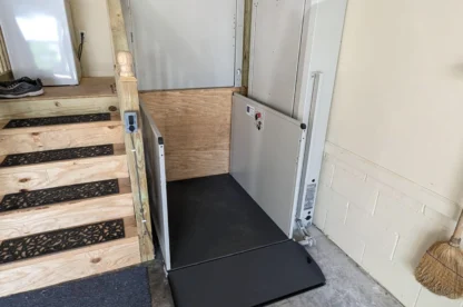A garage with a wooden staircase to the left and a residential vertical platform lift installed next to it. A broom is leaning against the wall on the right.