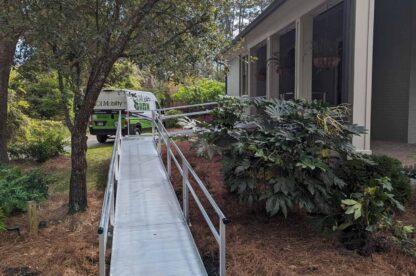 A metal accessibility ramp leads from a house to a pathway. A green van is parked nearby. Trees and shrubs surround the area.