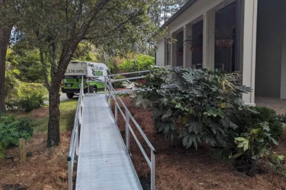 A metal accessibility ramp leads from a house to a pathway. A green van is parked nearby. Trees and shrubs surround the area.