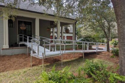 A house with a wheelchair ramp made of metal and wood, providing access to the front porch. The setup is surrounded by trees and bushes.