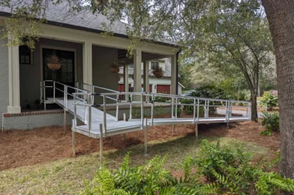 A house with a wheelchair ramp made of metal and wood, providing access to the front porch. The setup is surrounded by trees and bushes.