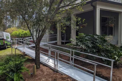 A metal wheelchair ramp leads from the driveway to the entrance of a house, surrounded by a garden with trees and plants.
