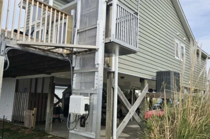 An exterior metal elevator shaft is attached to a raised house with a covered patio underneath. Steps lead up to the main floor next to the elevator.