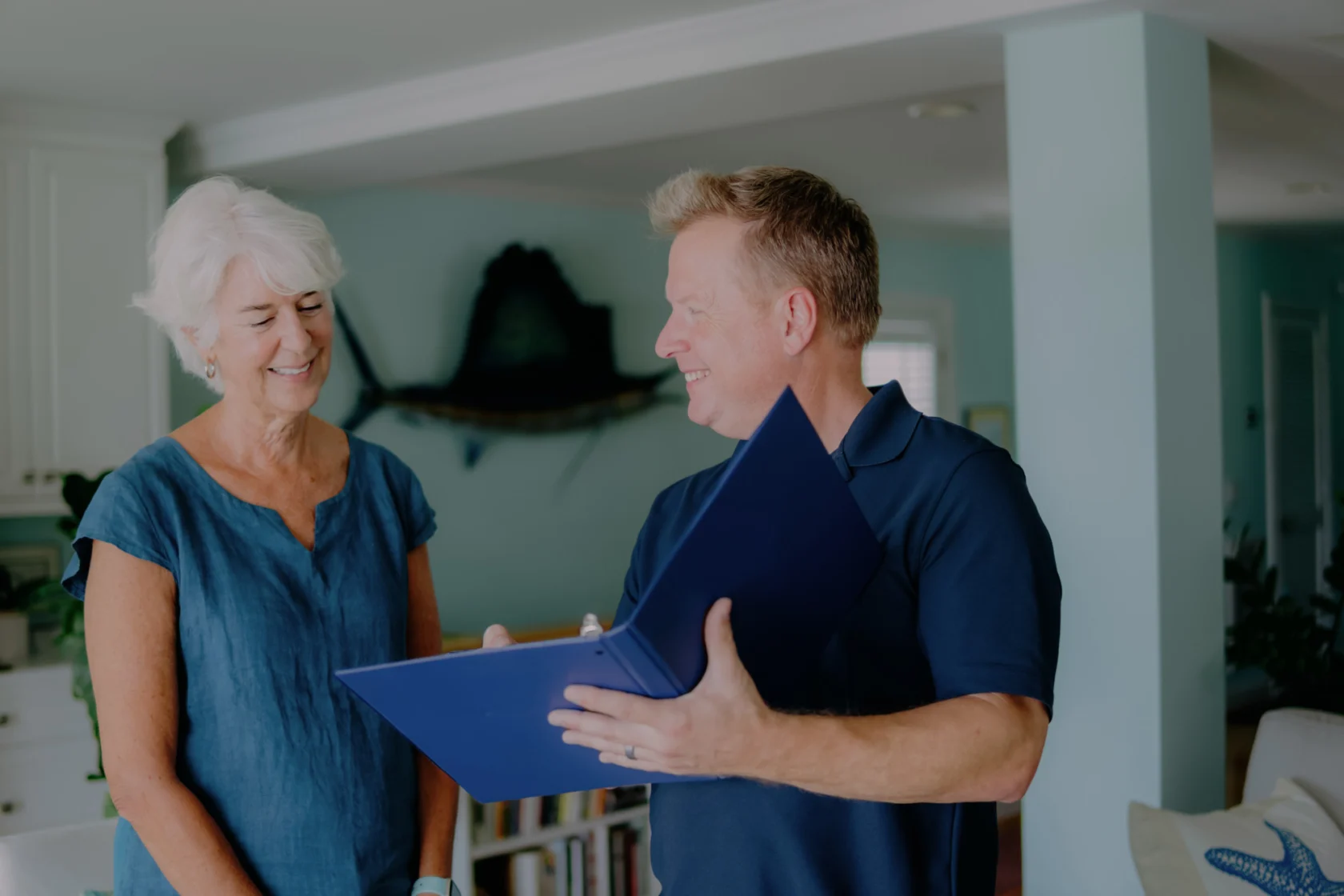 A man in a blue shirt holds an open folder and talks to an older woman in a blue top, standing together in a brightly lit room.