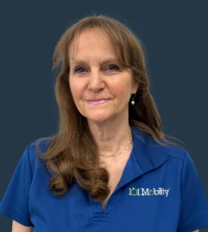 A woman with long brown hair wearing a blue shirt looks at the camera against a plain dark background.