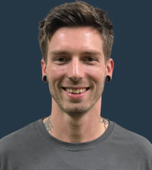 A person with short hair and ear gauges smiles, wearing a gray shirt and standing against a dark blue background.