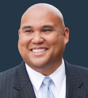 A bald man in a pinstripe suit jacket, white shirt, and light blue tie is smiling against a dark blue background.