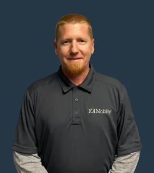 A man with short red hair and a goatee, wearing a gray "IOU Mobility" polo shirt stands against a solid dark background.