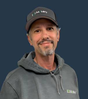 A man with a gray beard and mustache wears a gray hoodie and a matching cap in front of a solid dark blue background.