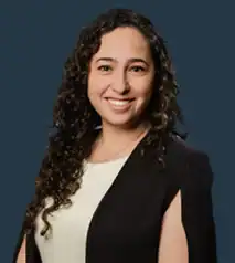 Woman with long curly hair wearing a white top and black blazer, smiling at the camera against a solid dark blue background.