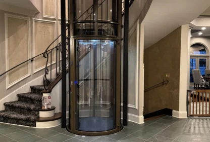 A glass cylinder elevator next to a curved staircase in an elegant foyer with tiled floors and decorative walls.