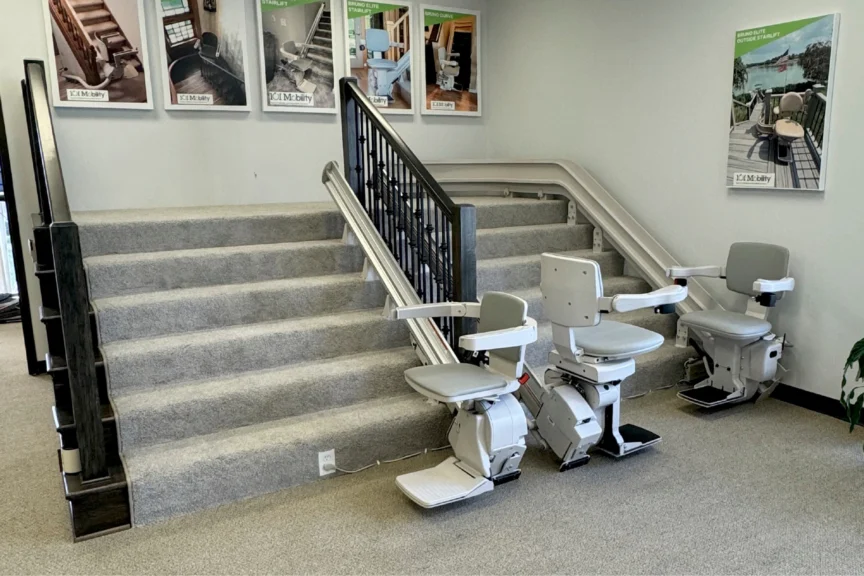 Showroom with grey-carpeted stairs featuring two stair lift chairs on rails. Posters of stair lift models are displayed on the wall.