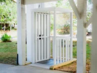 The Coastal Lift by Staying Home is a white outdoor elevator with glass sides and a ramp, nestled under a shelter, surrounded by lush grass and trees.