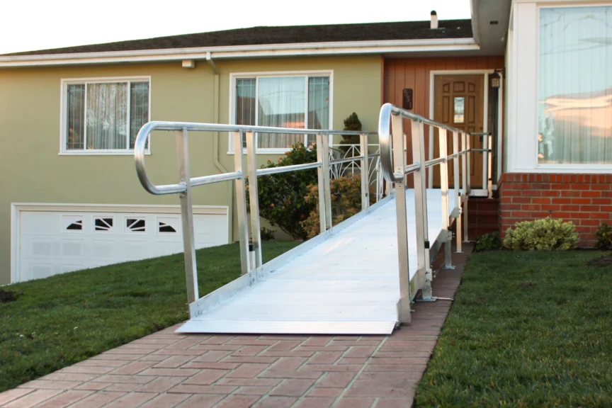 Aluminum wheelchair ramp leading to the front door of a single-story house with a green exterior and brick steps.