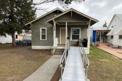 Single-story house with a ramp leading to the front porch, beside a lawn with trees and a row of bicycles on the left. A blue building with an "Open" sign is visible on the right.
