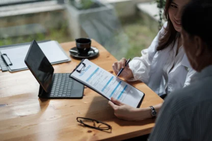 Two people seated at a table discussing a document on a clipboard, with a laptop, cup, and eyeglasses nearby.