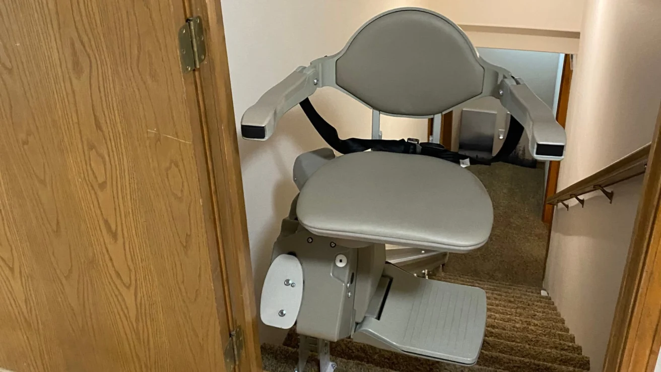 A gray stair lift chair installed on a staircase with beige carpeting and wooden side paneling.
