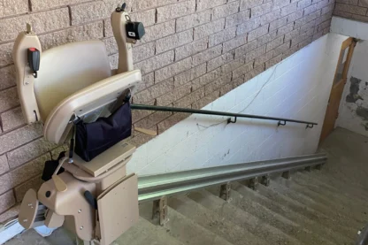 A stairlift attached to a brick wall beside a staircase, with a safety rail on the opposite side.