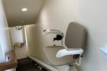A stairlift is installed on a residential staircase, featuring a beige seat and armrests, with a remote control attached.