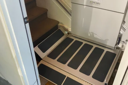 A stairlift platform at the base of a staircase, with a folded seat and safety barriers visible.